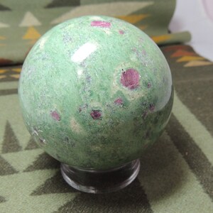May include: A green and pink sphere with a smooth, polished surface. The sphere is made of a type of gemstone with pink inclusions. It is displayed on a clear stand.