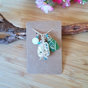 May include: A silver necklace featuring a pendant with a starfish, sea glass, and a white floral charm. The pendant is wire-wrapped with blue beads. The necklace is displayed on a brown card.