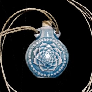 May include: A light blue ceramic pendant with a white floral design and a cork stopper. The pendant is suspended from a beige cord. The design is a mandala-like pattern. The background is black.