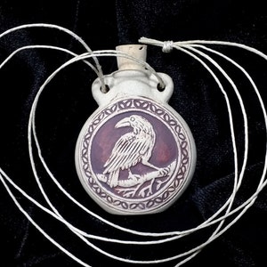 May include: A white ceramic pendant with a raven design, featuring a raven perched on a branch within a circular frame. The pendant has a cork stopper and is strung on a white cord. The background is black.