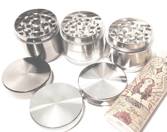 Puede incluir: Tres molinillos de metal plateado con una tapa de metal plateado y una base de metal plateado. Los molinillos tienen un diseño circular con una serie de agujeros. Los molinillos están sobre una superficie blanca. Un encendedor con un diseño marrón y blanco también está sobre la superficie.