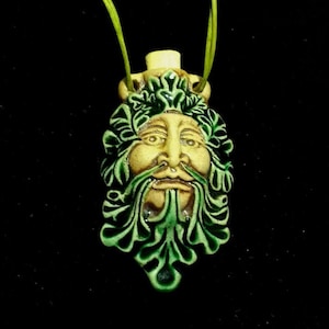 May include: A pendant featuring a Green Man face with a light green cord. The face has a tan complexion, green beard, and leafy crown. The pendant is a unique piece of jewelry.