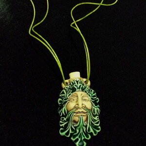 May include: A ceramic pendant featuring a Green Man face with a cream-colored face, green beard, and green leafy details. The pendant is strung on a lime green cord. The background is black.