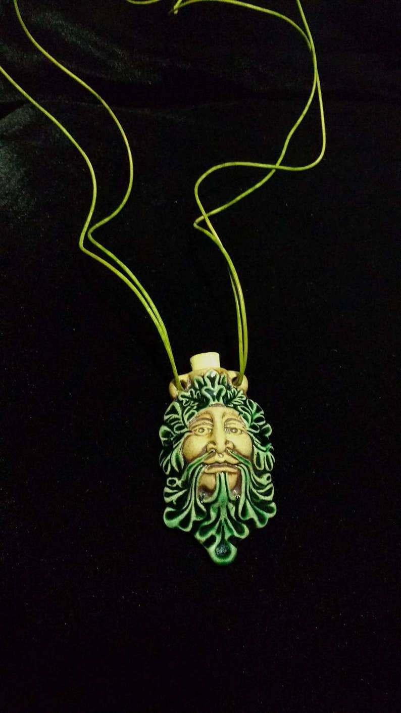 May include: A ceramic pendant featuring a Green Man face with a cream-colored face, green beard, and green leafy details. The pendant is strung on a lime green cord. The background is black.