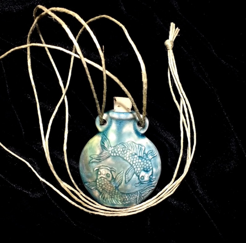 May include: A blue ceramic pendant with two fish carved into the surface. The pendant is hanging from a twine cord.