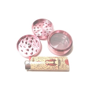 K&ouml;nnte beinhalten: Ein rosa Metall-Grinder mit drei Teilen, darunter ein Oberteil mit L&ouml;chern, ein Unterteil mit einem Sieb und ein Mittelteil mit Z&auml;hnen. Ein Feuerzeug mit einem Meerjungfrauen-Motiv ist ebenfalls enthalten.