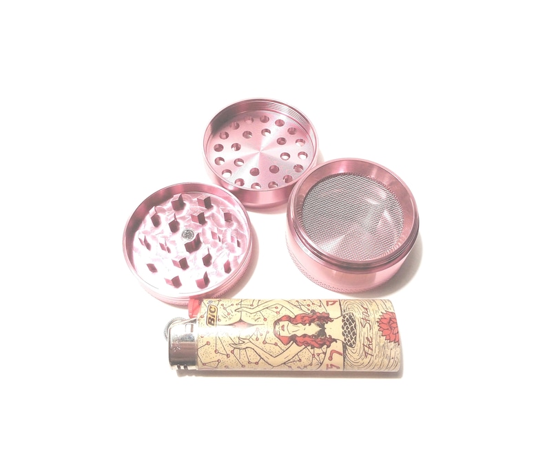 K&ouml;nnte beinhalten: Ein rosa Metall-Grinder mit drei Teilen, darunter ein Oberteil mit L&ouml;chern, ein Unterteil mit einem Sieb und ein Mittelteil mit Z&auml;hnen. Ein Feuerzeug mit einem Meerjungfrauen-Motiv ist ebenfalls enthalten.