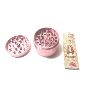 K&ouml;nnte beinhalten: Ein rosa Metall-Grinder mit abnehmbarem Deckel und ein Feuerzeug mit einem Meerjungfrauen-Motiv und dem Text "The Star".