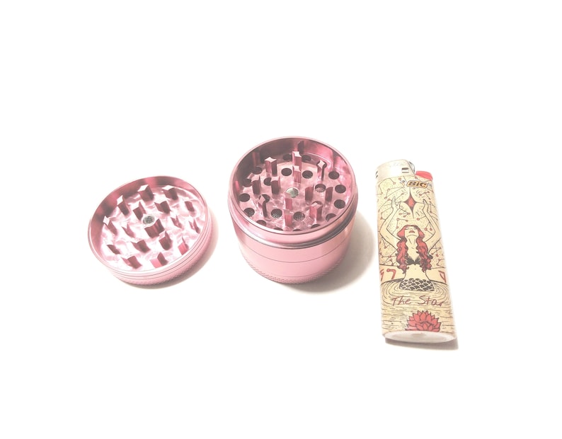 K&ouml;nnte beinhalten: Ein rosa Metall-Grinder mit abnehmbarem Deckel und ein Feuerzeug mit einem Meerjungfrauen-Motiv und dem Text "The Star".