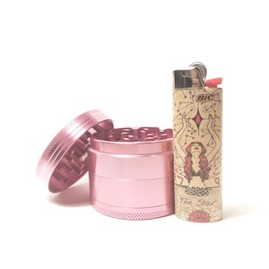 K&ouml;nnte beinhalten: Ein rosa Metall-Grinder mit einem separaten Deckel und einem Feuerzeug mit einem Meerjungfrauen-Design und dem Text "The Star" und "BIC".