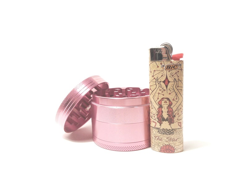 K&ouml;nnte beinhalten: Ein rosa Metall-Grinder mit einem separaten Deckel und einem Feuerzeug mit einem Meerjungfrauen-Design und dem Text "The Star" und "BIC".