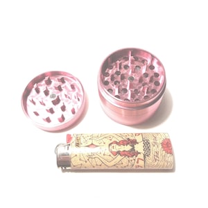 K&ouml;nnte beinhalten: Ein rosa Metall-Grinder mit einem separaten Deckel und einem Feuerzeug mit einem Meerjungfrauen-Motiv. Das Feuerzeug hat eine Standardgr&ouml;&szlig;e und ein goldenes BIC-Logo.