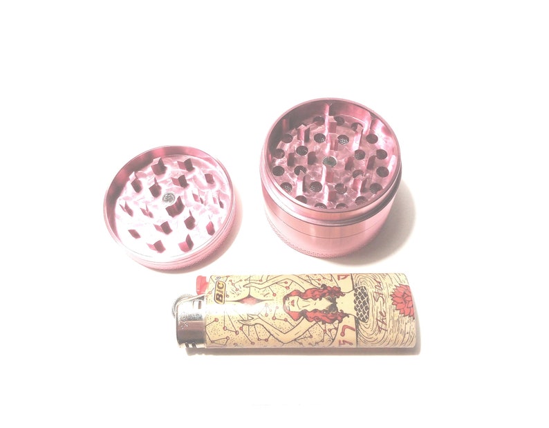 K&ouml;nnte beinhalten: Ein rosa Metall-Grinder mit einem separaten Deckel und einem Feuerzeug mit einem Meerjungfrauen-Motiv. Das Feuerzeug hat eine Standardgr&ouml;&szlig;e und ein goldenes BIC-Logo.