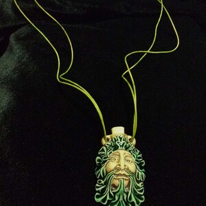 May include: A ceramic pendant necklace featuring a Green Man face with a beard and leafy details. The pendant is cream, green, and brown, suspended from a lime green cord. The necklace is displayed against a black background.