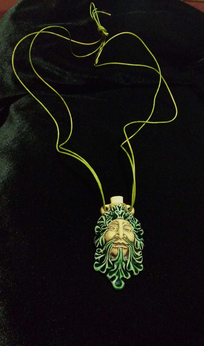 May include: A ceramic pendant necklace featuring a Green Man face with a beard and leafy details. The pendant is cream, green, and brown, suspended from a lime green cord. The necklace is displayed against a black background.