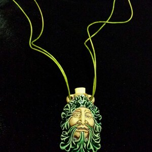 May include: A ceramic pendant necklace featuring a Green Man face with a green leafy beard and crown. The face is tan with green accents. The necklace has a lime green cord. The background is black.