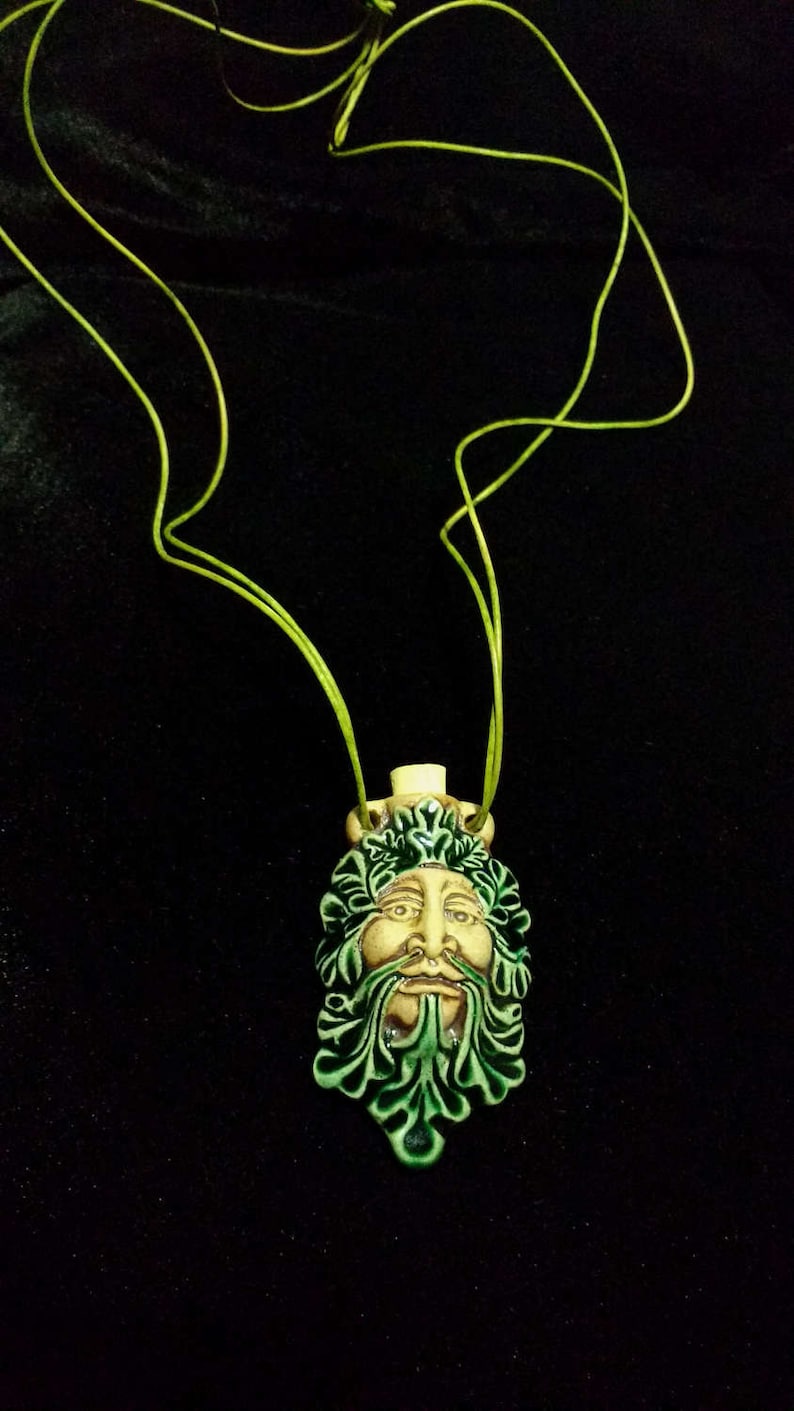 May include: A ceramic pendant necklace featuring a Green Man face with a green leafy beard and crown. The face is tan with green accents. The necklace has a lime green cord. The background is black.