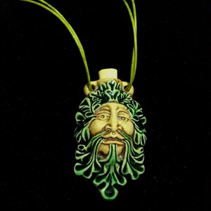 May include: A ceramic pendant featuring a Green Man face with green leaf details and a cream-colored face. The pendant hangs from a green cord against a black background. The pendant is approximately 2 inches in height.