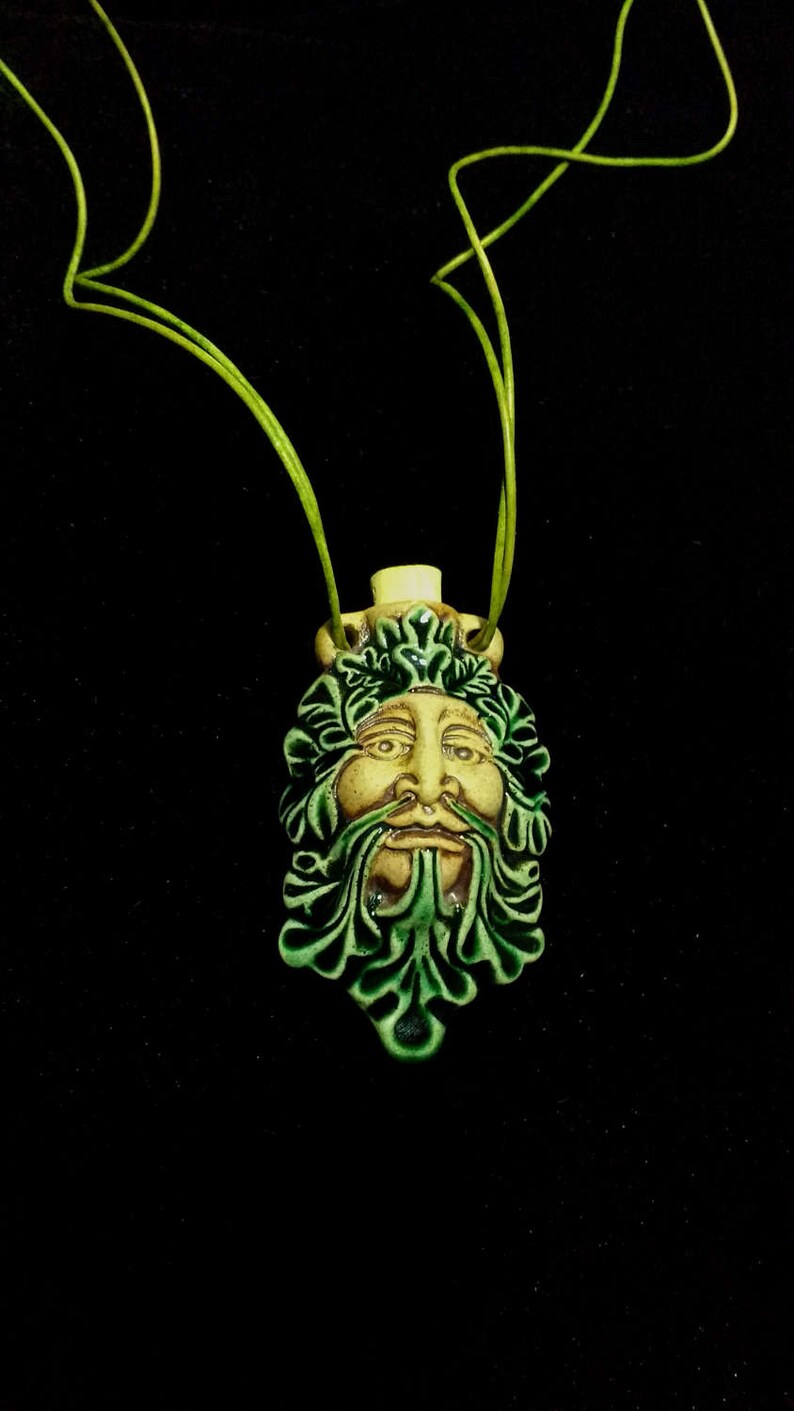 May include: A ceramic pendant featuring a Green Man face with green leaf details and a cream-colored face. The pendant hangs from a green cord against a black background. The pendant is approximately 2 inches in height.