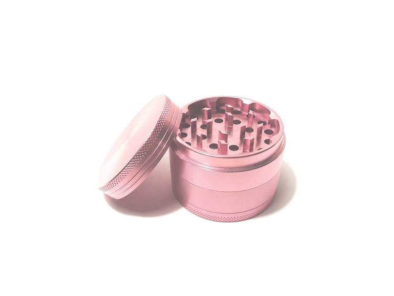 K&ouml;nnte beinhalten: Ein rosa Metall-Grinder mit vier Abschnitten. Der obere Abschnitt hat eine Reihe von L&ouml;chern zum Mahlen von Kr&auml;utern. Der Grinder ist offen und der obere Abschnitt ist abgenommen.