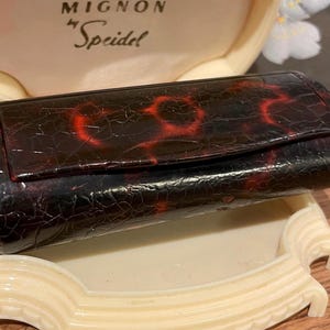 May include: A vintage, rectangular, dark brown case with a red, crackled pattern. The case has a flap closure and is resting on a cream-colored, ornate tray. The text "MIGNON by Speidel" is visible in the background.
