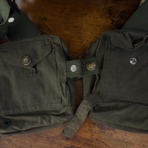 May include: An olive green canvas chest pack with two front pockets, secured by buttons. The pack features adjustable straps and is designed to be worn across the chest. The buttons are embossed with text.