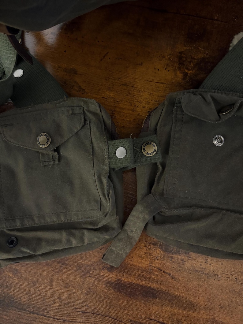 May include: An olive green canvas chest pack with two front pockets, secured by buttons. The pack features adjustable straps and is designed to be worn across the chest. The buttons are embossed with text.