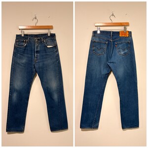May include: A pair of blue denim jeans shown from the front and back. The front features a button fly and five-pocket construction. The back displays two pockets with arcuate stitching, a red tab, and a tan leather patch on the waistband.