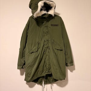Puede incluir: Parka militar verde oliva con capucha forrada de piel sintética blanca. La parka presenta un parche de "U.S. ARMY", dos bolsillos delanteros y cintura con cordón. Una prenda de abrigo clásica y duradera.