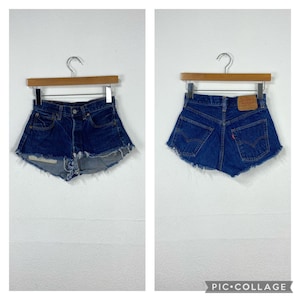 Puede incluir: Un par de pantalones cortos de mezclilla azul con bordes deshilachados. Los pantalones cortos tienen cintura alta y se muestran colgados en una percha de madera.