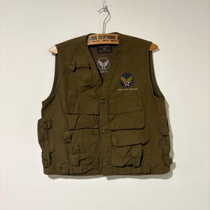 May include: Olive green vest with multiple pockets and button fastenings. Features two patches: one with a winged emblem and the text "AIR FO", and another with a star and the text "ARMY AIR FORCES". The vest is hanging on a wooden hanger.