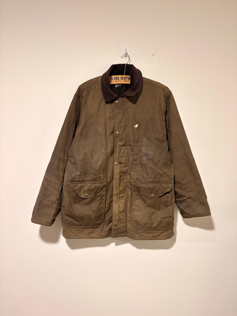 Peut inclure: Veste en coton cir&eacute; marron avec un col en velours c&ocirc;tel&eacute; et deux grandes poches avant. La veste a une fermeture boutonn&eacute;e et un petit embl&egrave;me brod&eacute; sur la poitrine. L'&eacute;tiquette indique "GLOBE CLOTH".