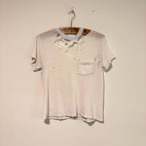 May include: A short-sleeved, off-white t-shirt featuring a crew neckline and a single chest pocket. The garment displays significant distressing, including multiple holes and frayed edges around the collar and upper chest area, hanging on a wooden hanger.