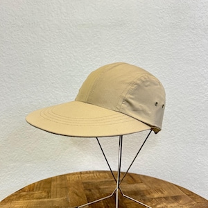 May include: A tan-colored cap with a wide brim, displayed on a metal stand. The cap has a curved visor and a structured crown. The hat is made of a lightweight fabric and features a simple, minimalist design. The hat is approximately 10 inches in diameter.