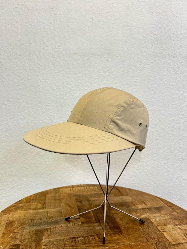 May include: A tan-colored cap with a wide brim, displayed on a metal stand. The cap has a curved visor and a structured crown. The hat is made of a lightweight fabric and features a simple, minimalist design. The hat is approximately 10 inches in diameter.