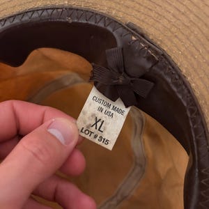 Pode incluir: Chap&eacute;u de palha com faixa e interior em couro castanho. Uma etiqueta dentro do chap&eacute;u diz "CUSTOM MADE IN USA" e "XL LOT #315". O chap&eacute;u tem uma aba larga e uma fita castanha escura.
