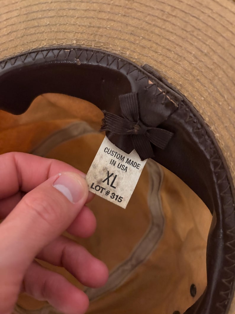 Pode incluir: Chap&eacute;u de palha com faixa e interior em couro castanho. Uma etiqueta dentro do chap&eacute;u diz "CUSTOM MADE IN USA" e "XL LOT #315". O chap&eacute;u tem uma aba larga e uma fita castanha escura.