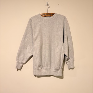 Peut inclure: Un sweat-shirt gris clair à col rond avec des manches longues à poignets côtelés et un ourlet côtelé. Le vêtement présente un petit logo Champion brodé en rouge, blanc et bleu sur la manche gauche près du poignet, suspendu à un cintre en bois.