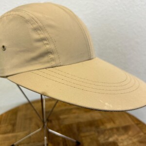 May include: A tan-colored cap with a wide brim. The cap has a structured crown with stitched seams and a small vent on the side. The brim has multiple stitched lines for reinforcement. The cap is displayed on a metal stand.