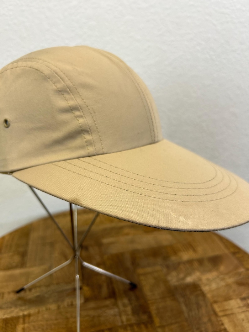 May include: A tan-colored cap with a wide brim. The cap has a structured crown with stitched seams and a small vent on the side. The brim has multiple stitched lines for reinforcement. The cap is displayed on a metal stand.