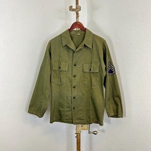 Può includere: Una camicia da lavoro verde a maniche lunghe con due tasche sul petto. La camicia ha una chiusura con bottoni sul davanti e una toppa sulla manica sinistra con un motivo a chevron.