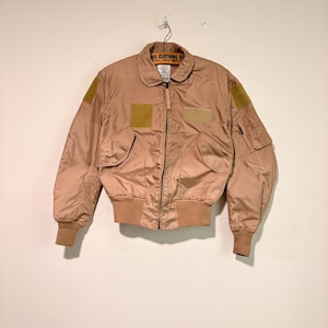 May include: A tan bomber jacket featuring a front zip fastening, ribbed cuffs and hem, two snap-button flap pockets, and a utility pocket on the left sleeve. The jacket includes rectangular hook-and-loop patches on the chest and shoulders.