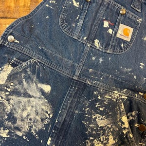 May include: Blue denim overalls with white paint splatters and a Carhartt logo patch. The overalls have multiple pockets and adjustable straps.