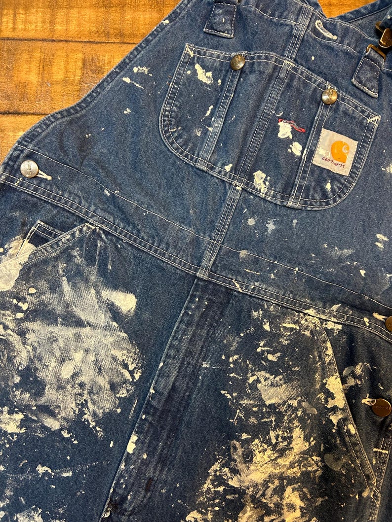 May include: Blue denim overalls with white paint splatters and a Carhartt logo patch. The overalls have multiple pockets and adjustable straps.