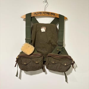 May include: Olive green fishing vest with two front pockets, a central pocket, and adjustable straps. The vest has a light brown sheepskin patch and is hanging on a wooden hanger with text.