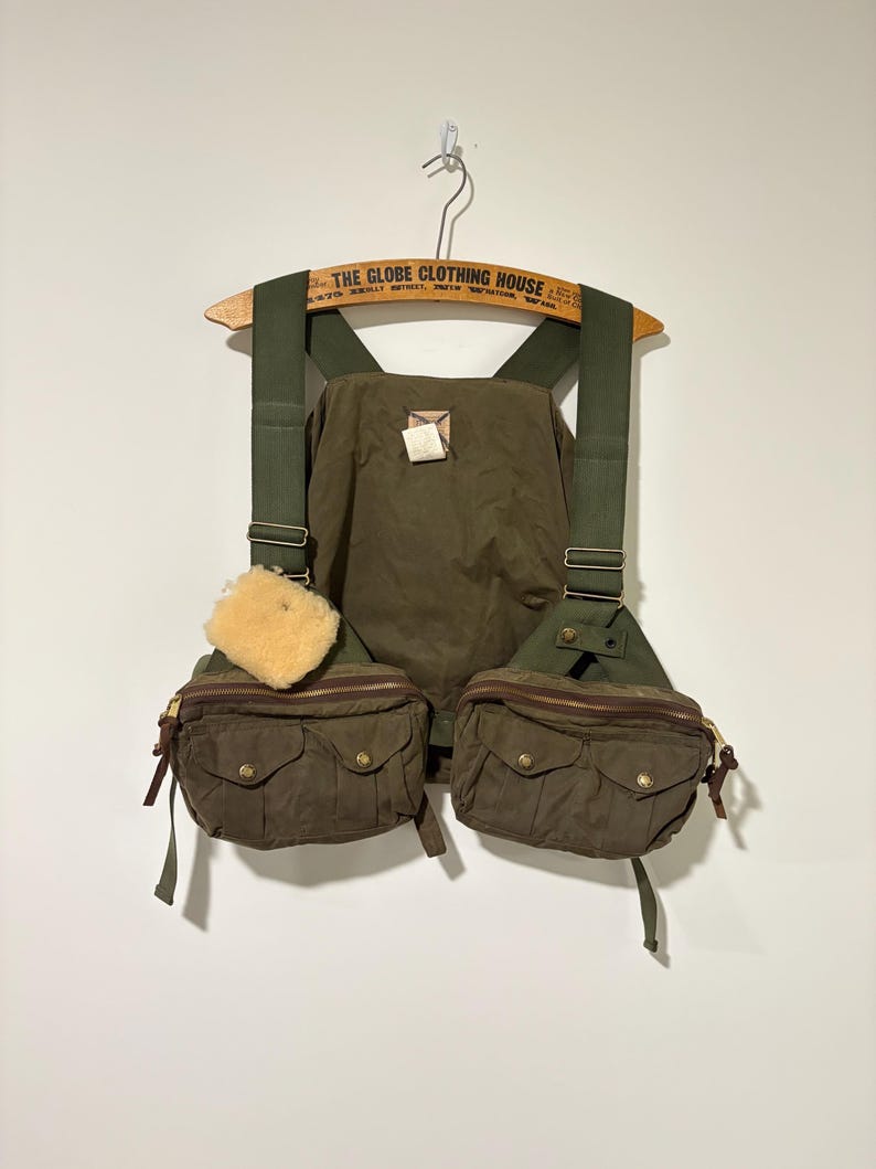 May include: Olive green fishing vest with two front pockets, a central pocket, and adjustable straps. The vest has a light brown sheepskin patch and is hanging on a wooden hanger with text.