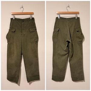 Può includere: Pantaloni cargo verde oliva con più tasche, appesi a una gruccia di legno. I pantaloni hanno una chiusura con bottoni e una vestibilità ampia. L'immagine mostra sia la parte anteriore che posteriore dei pantaloni.