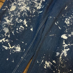May include: A pair of dark blue denim jeans with white paint splatters all over them. The jeans have a seam down the side of one leg.