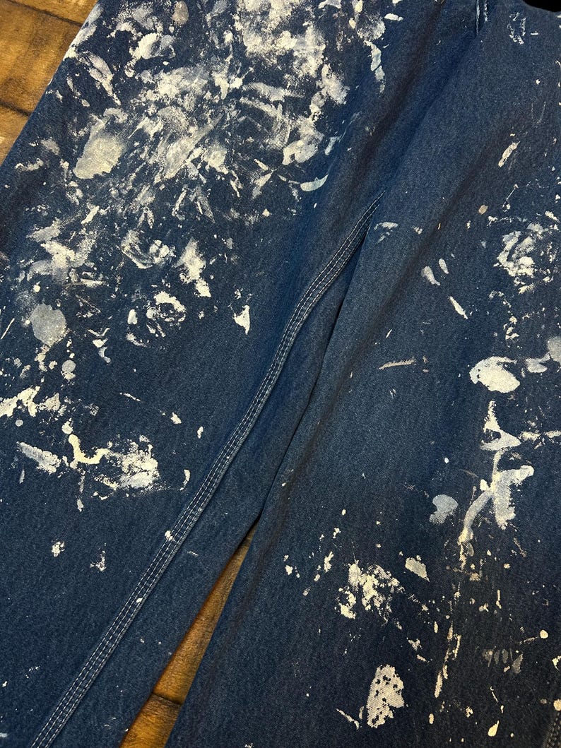May include: A pair of dark blue denim jeans with white paint splatters all over them. The jeans have a seam down the side of one leg.
