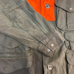 May include: A brown and orange waxed cotton hunting jacket with multiple pockets and button closures. The jacket has a  "Filson" logo on the sleeve.